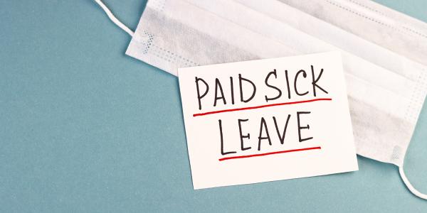 a face mask and piece of white paper with the words 'PAID SICK LEAVE' written on it  against a blue background. 