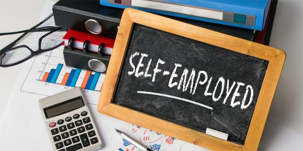 a desk with a calculator, glasses, folders, and a chalk board with the word 'SELF-EMPLOYED' written on it. a desk with a calculator, glasses, folders, and a chalk board with the word 'SELF-EMPLOYED' written on it.