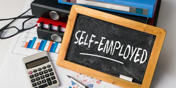 a desk with a calculator, glasses, folders, and a chalk board with the word 'SELF-EMPLOYED' written on it. 