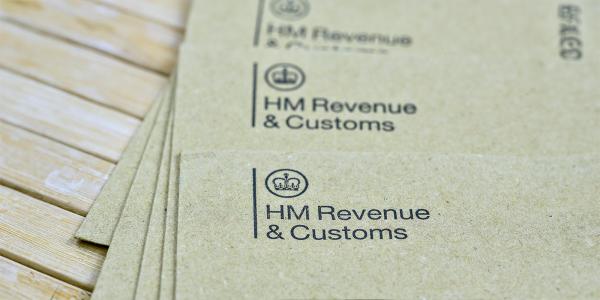 a pile of brown envelopes on a wooden table, each envelope has 'HM REVENUE & CUSTOMS' printed in the top left corner along with the HMRC logo. 
