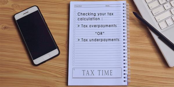 pad of paper on a desk next to a keyboard and mobile phone, pad of paper reads 'CHECKING YOUR TAX CALCULATION: TAX OVERPAYMENTS OR TAX UNDERPAYMENTS'