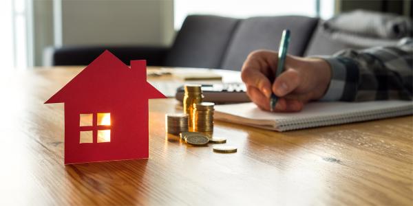 A person sat at a desk writing, also on the desk is a small red house and a stack of coins. 