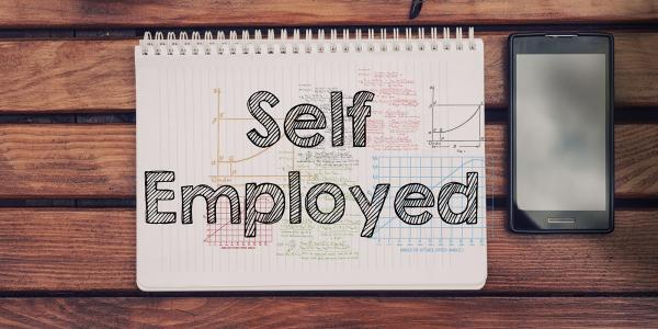 a wooden table with a mobile phone and a pad of paper with the words 'SELF EMPLOYMENT' written on it. 