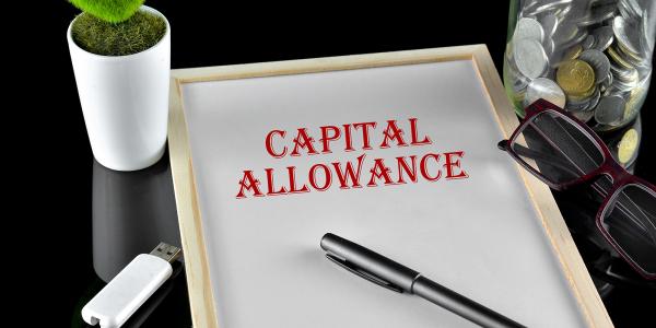 a black table top with a drawing board, on the board the words 'CAPITAL ALLOWANCE' are written in red pen. Next to this is a USB stick, a plant, some glasses, and a jar of coins a black table top with a drawing board, on the board the words 'CAPITAL ALLOWANCE' are written in red pen. Next to this is a USB stick, a plant, some glasses, and a jar of coins