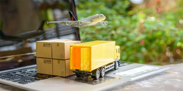 A laptop with tiny cardboard boxes and a toy haulage truck sat on the keyboard, a toy aeroplane is flying over the top of those.