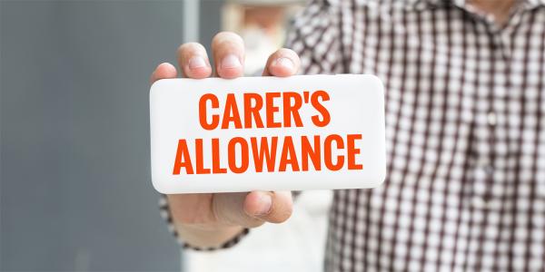 a person holding up a white card that says 'CARER'S ALLOWANCE' in orange text