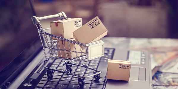 an open laptop with a tiny shopping trolley sat on the keyboard, inside the trolley are tiny brown parcels. 