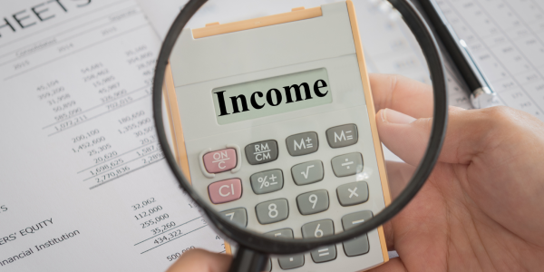 a person holding a magnifying glass up to a calculator showing the word 'INCOME' on the screen, behind this paperwork can be seen.