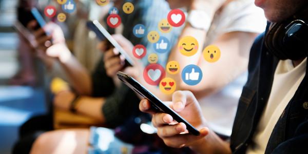 people looking at social media on their mobile phones, surrounded by social media reaction icons such as thumbs up, heart, smiley face. 