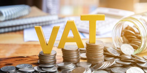 A desk scattered with coins and notebooks, a glass jar of coins is tipped on its side, three piles of coins can be seen in the centre each with a yellow letter on top. Together this spells 'VAT'.