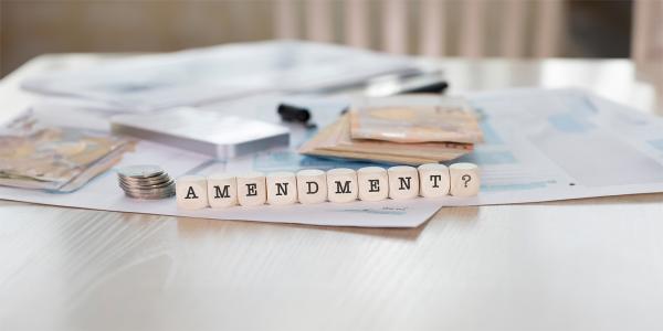 a desk with money and paperwork in the background with wooden blocks spelling out the word 'AMENDMENT'