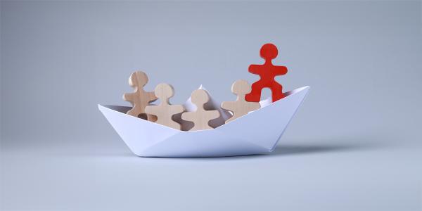 a paper boat with small wooden people inside, the person at the front of the boat is coloured red. 
