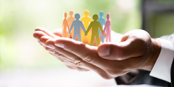 A person holding a paper chain of pastel coloured people holding hands stood in a circle. people are coloured in Pink, Green, Blue, Orange, Yellow. 