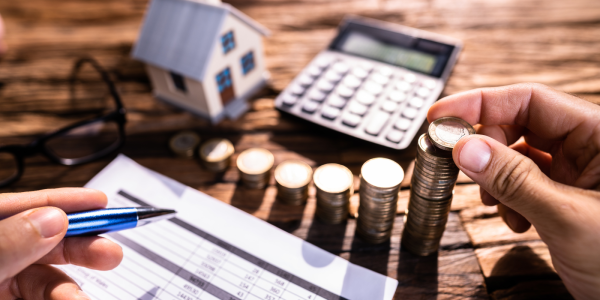 A person at a desk holding a pen with paperwork, stacks of coins, a small toy house and a calculator. A person at a desk holding a pen with paperwork, stacks of coins, a small toy house and a calculator.
