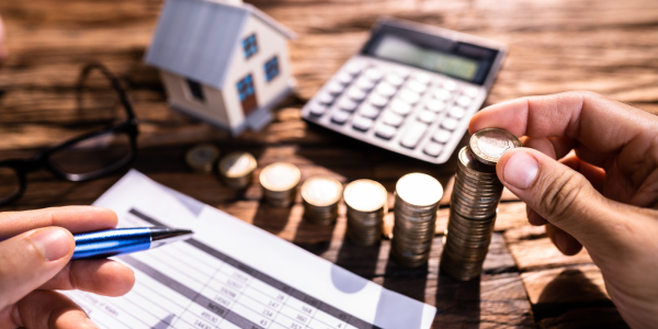 A person at a desk holding a pen with paperwork, stacks of coins, a small toy house and a calculator.