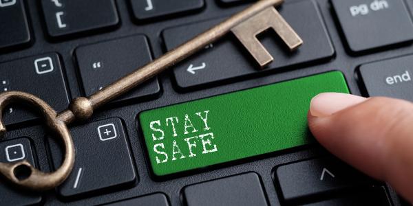 A keyboard with a Green key, a person is pressing this key. The key has white text on it that reads 'STAY SAFE' an old fashioned brass coloured key is placed on top of the keyboard. 