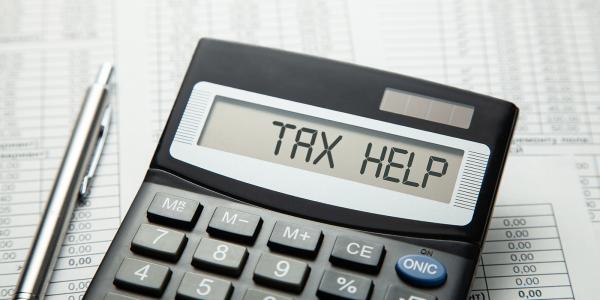 A calculator with the words 'TAX HELP' on the screen, next to the calculator is a pen, the background is made up of paperwork. A calculator with the words 'TAX HELP' on the screen, next to the calculator is a pen, the background is made up of paperwork.