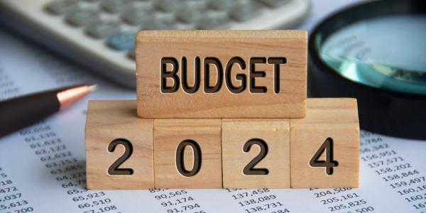 stacked wooden blocks, each block has engraved writing in black text on the side that together reads 'BUDGET 2024' background briefly shows a calculator, magnifying glass, a pen and paperwork. 