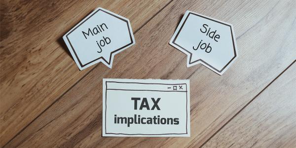 3 pieces of paper on a wooden desktop, one says 'TAX IMPLICATIONS', the other two are shaped like speech bubbles one saying 'MAIN JOB'. the other 'SIDE JOB' 