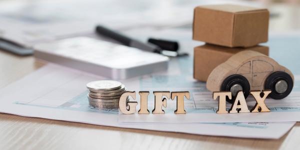 a desk showing paperwork, coins, a phone and a wooden car with wooden letters that spell out 'GIFT TAX' a desk showing paperwork, coins, a phone and a wooden car with wooden letters that spell out 'GIFT TAX'