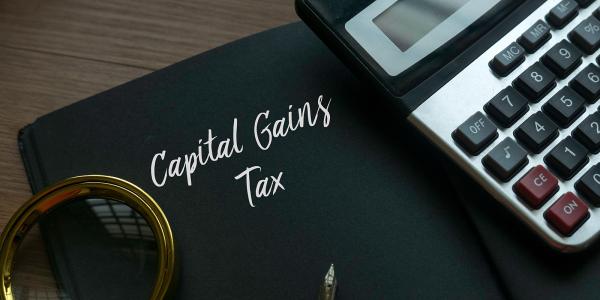 a black book with the words 'CAPITAL GAINS TAX' on the front. next to this is a calculator, a fountain pen and a magnifying glass. a black book with the words 'CAPITAL GAINS TAX' on the front. next to this is a calculator, a fountain pen and a magnifying glass.