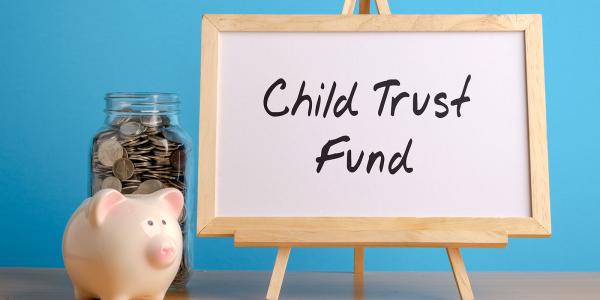 a wooden easel with the words 'CHILD TRUST FUND' written in black pen, next to this a piggy bank and a jar of coins can be seen against a blue background. a wooden easel with the words 'CHILD TRUST FUND' written in black pen, next to this a piggy bank and a jar of coins can be seen against a blue background.