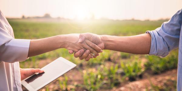 2 people shaking hands, one is holding a tablet in their other hand. The background is of countryside. 2 people shaking hands, one is holding a tablet in their other hand. The background is of countryside.