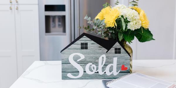 a bunch of flowers and a model house with the word 'SOLD' on the front sat on a kitchen countertop.