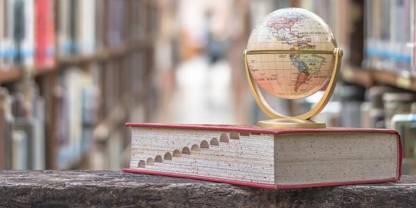 a large textbook with a globe of the world sat on top. background shows a library with many shelves of books. a large textbook with a globe of the world sat on top. background shows a library with many shelves of books.