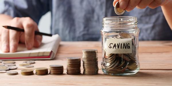 a person with a notebook and pen sat at a table with stacks of coins, the person is putting coins into a glass jar marked 'SAVING' a person with a notebook and pen sat at a table with stacks of coins, the person is putting coins into a glass jar marked 'SAVING'