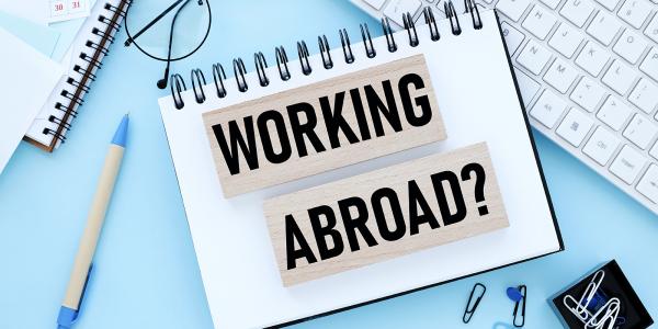 Two wooden blocks with the words 'WORKING ABROAD' showing on them in black text sat on top of a notebook on a persons desk. Scattered around the notebook various stationary can be seen and a computer keyboard. Two wooden blocks with the words 'WORKING ABROAD' showing on them in black text sat on top of a notebook on a persons desk. Scattered around the notebook various stationary can be seen and a computer keyboard.
