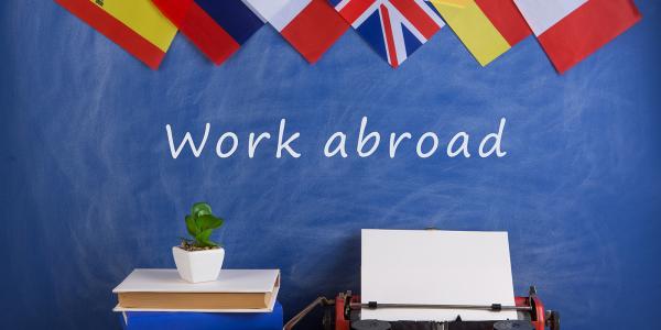 various flags can be seen at the top of the screen, below are a stack of books a small plant and a typewriter, on the wall the words 'WORK ABROAD' can be seen in white text various flags can be seen at the top of the screen, below are a stack of books a small plant and a typewriter, on the wall the words 'WORK ABROAD' can be seen in white text