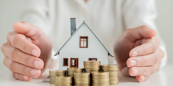 A small model house with stacks of coins outside, a person is cupping their hands on either side of the house. 