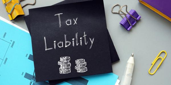 Black sheet of paper with the words 'TAX LIABILITY' written in white with an illustration of 2 piles of coins, various stationary surrounds the piece of black paper. Black sheet of paper with the words 'TAX LIABILITY' written in white with an illustration of 2 piles of coins, various stationary surrounds the piece of black paper.