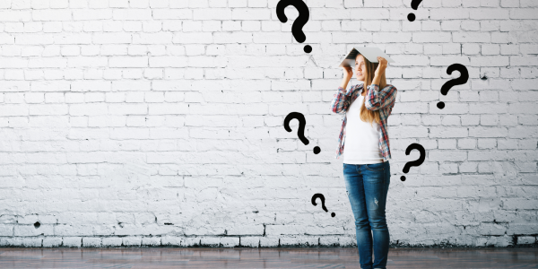 A person holding a book on their head, the white brick wall in the background has black question marks painted on it. A person holding a book on their head, the white brick wall in the background has black question marks painted on it.