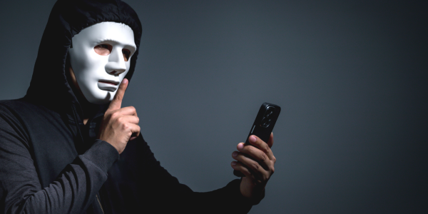 A person wearing all black with a white mask making the 'SSHHH' sign with their hand, in the other hand they are holding a phone. A person wearing all black with a white mask making the 'SSHHH' sign with their hand, in the other hand they are holding a phone.
