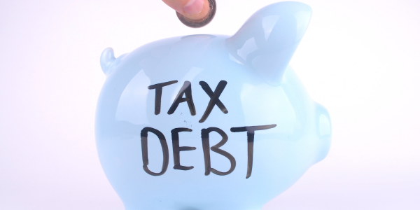 A person putting coins into a light coloured piggy bank with the words 'TAX DEBT' written in black ink. A person putting coins into a light coloured piggy bank with the words 'TAX DEBT' written in black ink.