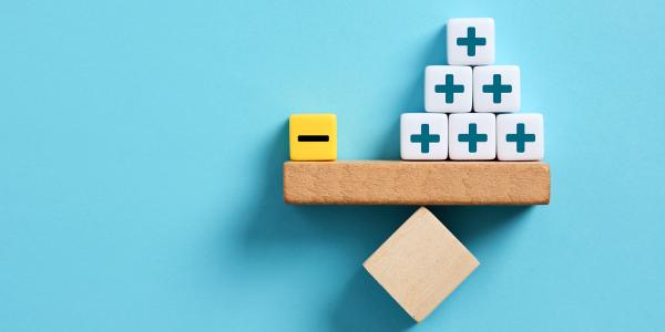 A wooden see saw perfectly balanced against a blue wall, on one side of the shelf is a yellow block with a minus sign on it, on the other side there are 9 white blocks with plus signs on each block. A wooden see saw perfectly balanced against a blue wall, on one side of the shelf is a yellow block with a minus sign on it, on the other side there are 9 white blocks with plus signs on each block.