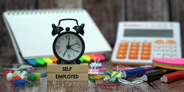 A desk scattered with various stationary, a clock and wooden blocks with the words 'SELF EPLOYED' printed on them in black text. A desk scattered with various stationary, a clock and wooden blocks with the words 'SELF EPLOYED' printed on them in black text.