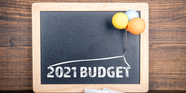 A chalkboard with the words '2021 BUDGET' printed on it, a small bunch o balloons are attached to the chalkboard. A chalkboard with the words '2021 BUDGET' printed on it, a small bunch o balloons are attached to the chalkboard.