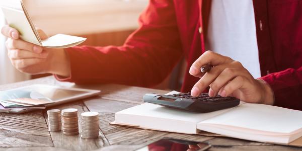 A person sat at a desk with paperwork and a calculator along with stacks of coins. A person sat at a desk with paperwork and a calculator along with stacks of coins.