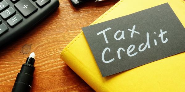 A black card showing the words 'TAX CREDIT' in white writing, the card is sat on top of a yellow notebook on a desk next to a computer keyboard. A black card showing the words 'TAX CREDIT' in white writing, the card is sat on top of a yellow notebook on a desk next to a computer keyboard.