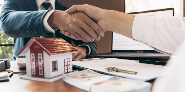 2 people shaking hands, on the desk sits a model home some cash, a key and paperwork. 2 people shaking hands, on the desk sits a model home some cash, a key and paperwork.