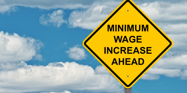 A blue sky with a scattering of white clouds and a yellow road sign with the words 'MINIMUM WAGE INCREASE AHEAD' in black text. A blue sky with a scattering of white clouds and a yellow road sign with the words 'MINIMUM WAGE INCREASE AHEAD' in black text.
