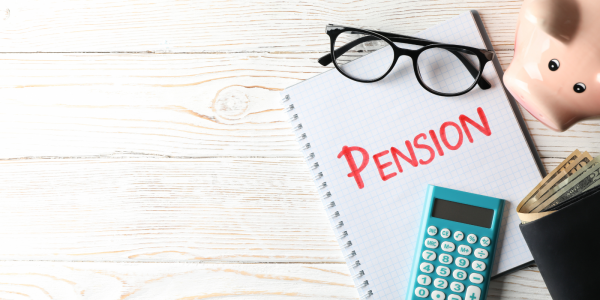 A desk with a piggy bank, a wallet,pair of glasses, a calculator and a notepad with the word 'PENSION' written on the front in red marker pen. A desk with a piggy bank, a wallet,pair of glasses, a calculator and a notepad with the word 'PENSION' written on the front in red marker pen.