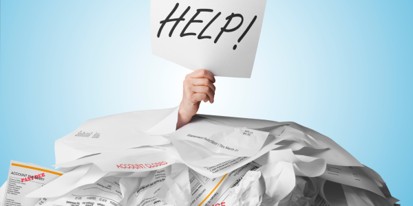 A person buried beneath a stack of overdue papers, a hand can be seen sticking out holding up a piece of paper with the word 'HELP!' written on it in black ink. A person buried beneath a stack of overdue papers, a hand can be seen sticking out holding up a piece of paper with the word 'HELP!' written on it in black ink.
