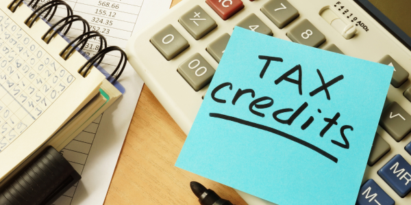 a blue post it note with the words 'TAX CREDITS' written on it in black pen, also on the desk is a notebook and a calculator. a blue post it note with the words 'TAX CREDITS' written on it in black pen, also on the desk is a notebook and a calculator.