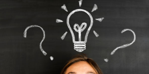 Person stood in front of a chalkboard, the chalkboard has a drawing of a lightbulb and question marks Person stood in front of a chalkboard, the chalkboard has a drawing of a lightbulb and question marks