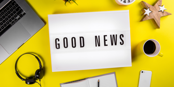 Light up box with the words 'GOOD NEWS' on it, a laptop, headphones, notepad and pen, coffee cup amongst other items are sat on the yellow desk.
