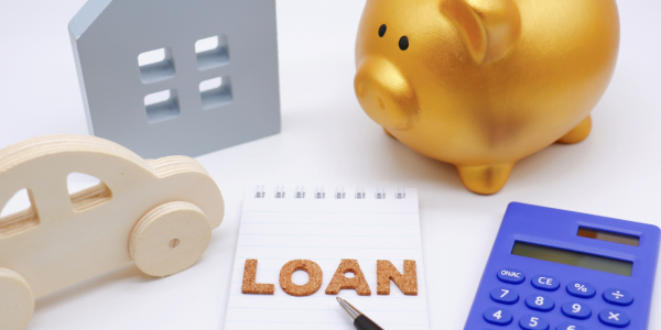 A white desk containing a golden piggy bank, a silver wooden house, a wooden car, the word 'LOAN' and a calculator. 